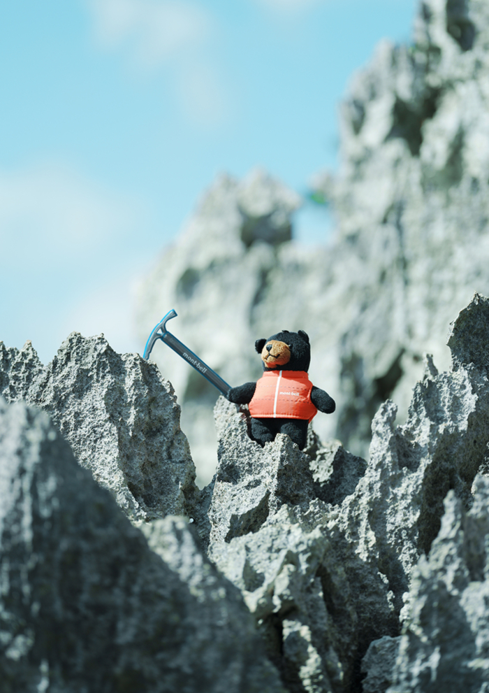 A Monta Bear plush toy wearing a red vest stands on a jagged rocky terrain, holding a small ice axe and gazing upwards into the distance.