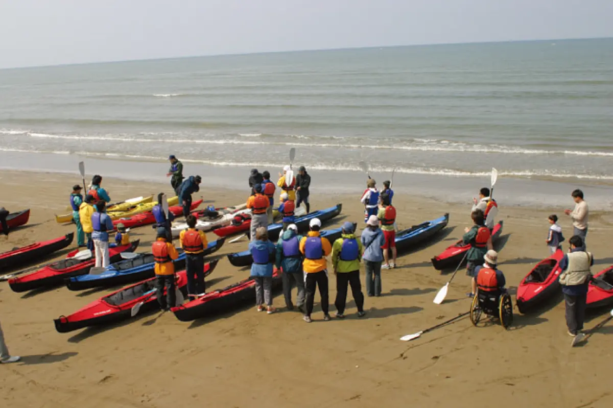 A scene from the outdoor event "Try & Carry" showing a kayaking experience session taking place on a beach. Many kayaks are lined up, and a large crowd has gathered.