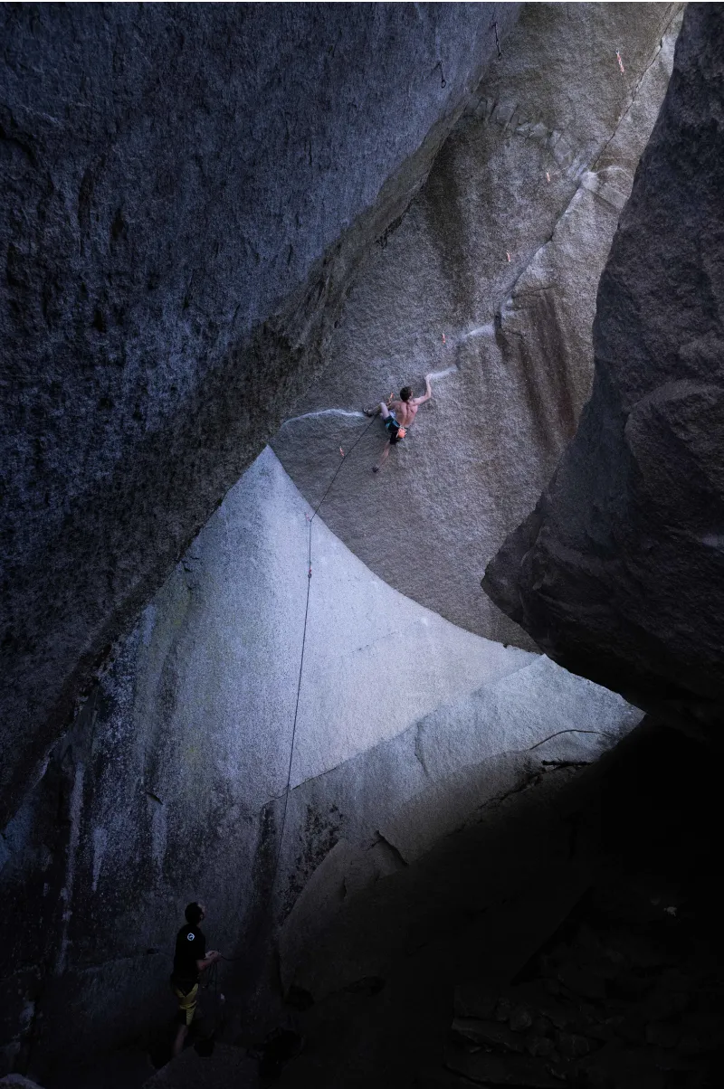A shirtless man with an orange chalk bag attached to his waist is climbing an overhanging dike, while a man in a black shirt is belaying him