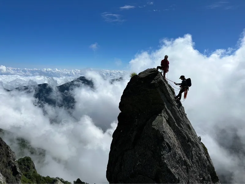 Two people, connected by a rope, are climbing a large rock while clouds are rising from below.