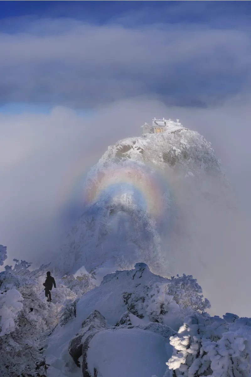 The summit of Mount Ishizuchi with its shrine is visible beyond a snowy ridgeline, with a double circular rainbow appearing in the foreground.