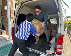 Mr. Noguchi in the back of a van, receiving cardboard boxes from a delivery staff member.