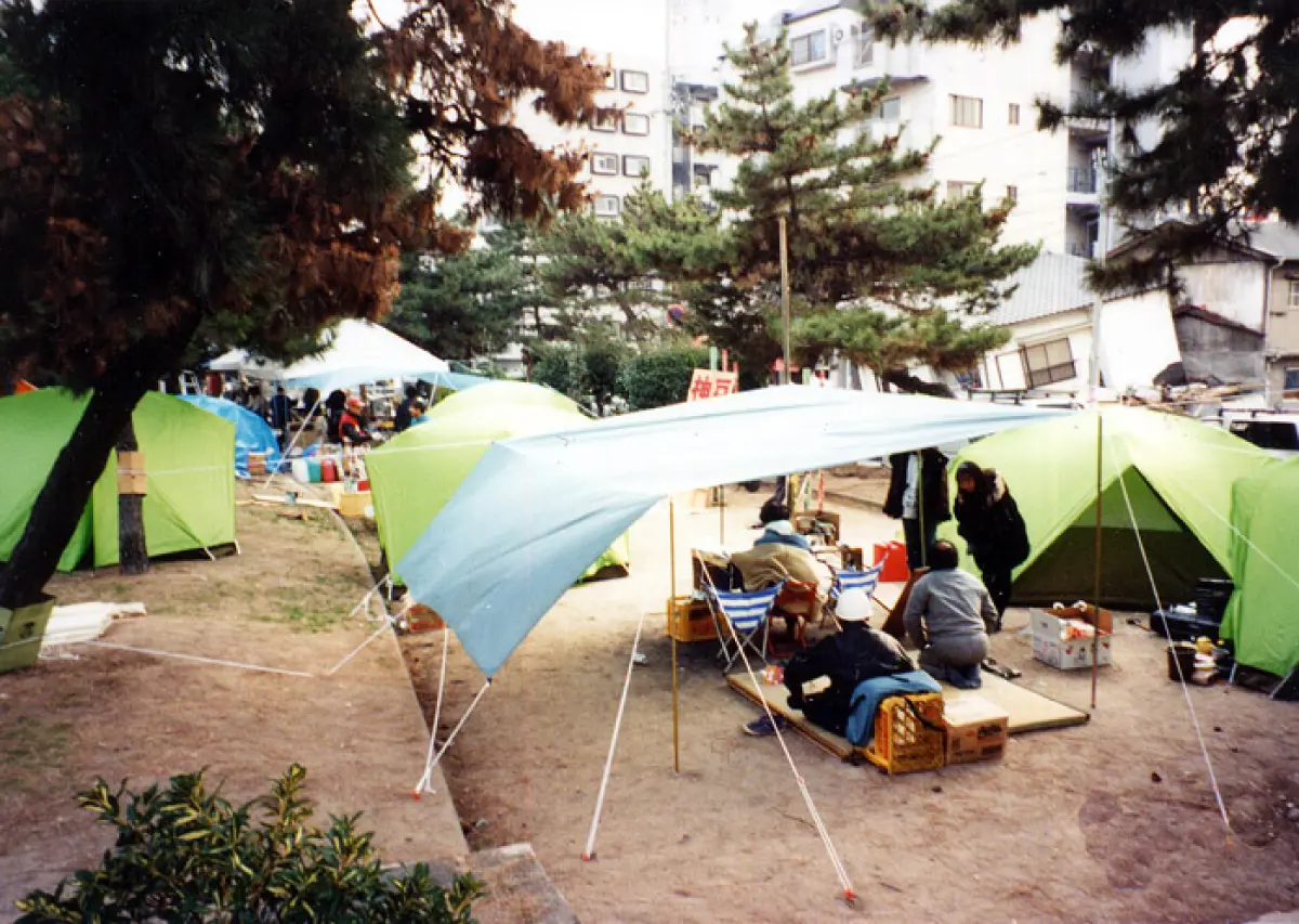 Scene of people living in tents set up in a park after the Great Hanshin-Awaji Earthquake. Collapsed houses remain in the surrounding area