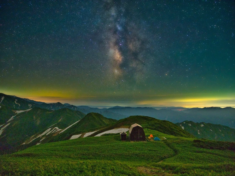 A landscape featuring a mountain hut and tents amidst expansive mountain ranges. The sky is filled with countless stars.