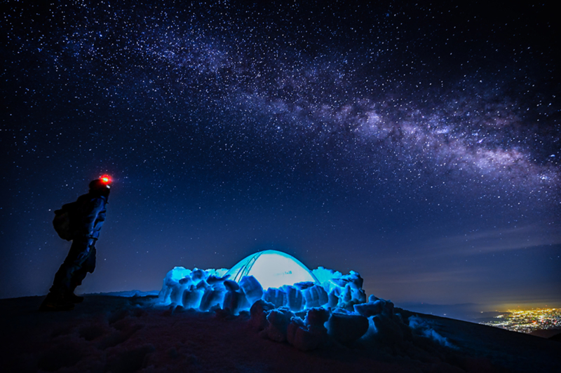 Beneath a sky filled with countless stars, a man with a red headlight stands alone, gazing upward. A tent, surrounded by snow walls, glows with a pale blue light from within, while in the distance, lights can be seen at the foot of the mountains.
