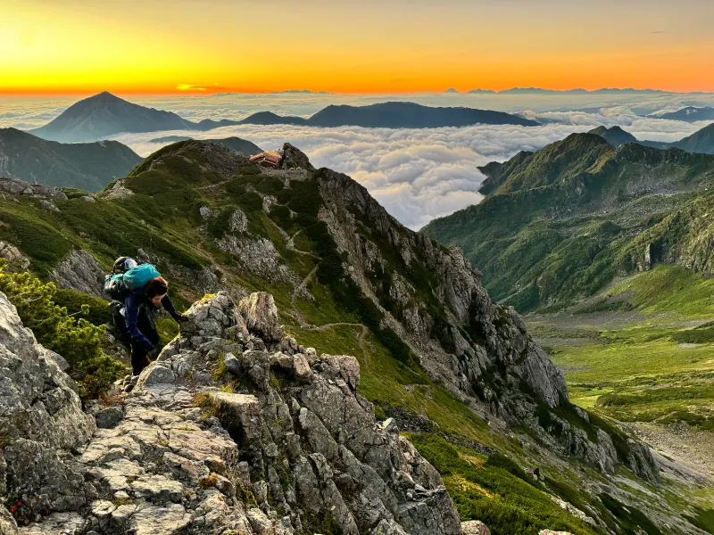 A woman is climbing rocks on a rugged ridgeline. Behind her, a sea of clouds spreads out, and the sky is dyed orange, suggesting that the sun is about to rise.