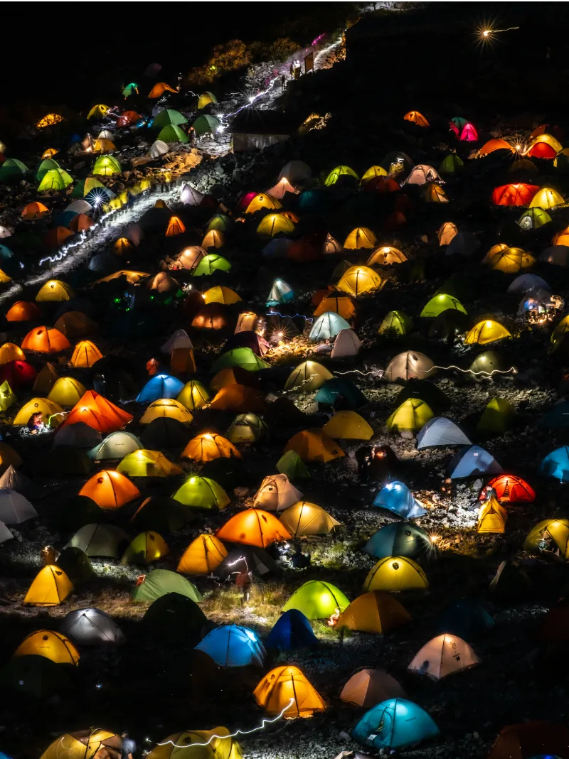 Colorful tents in yellow, green, blue, and other hues are lined up, with lights from mountaineers glowing in each of them.