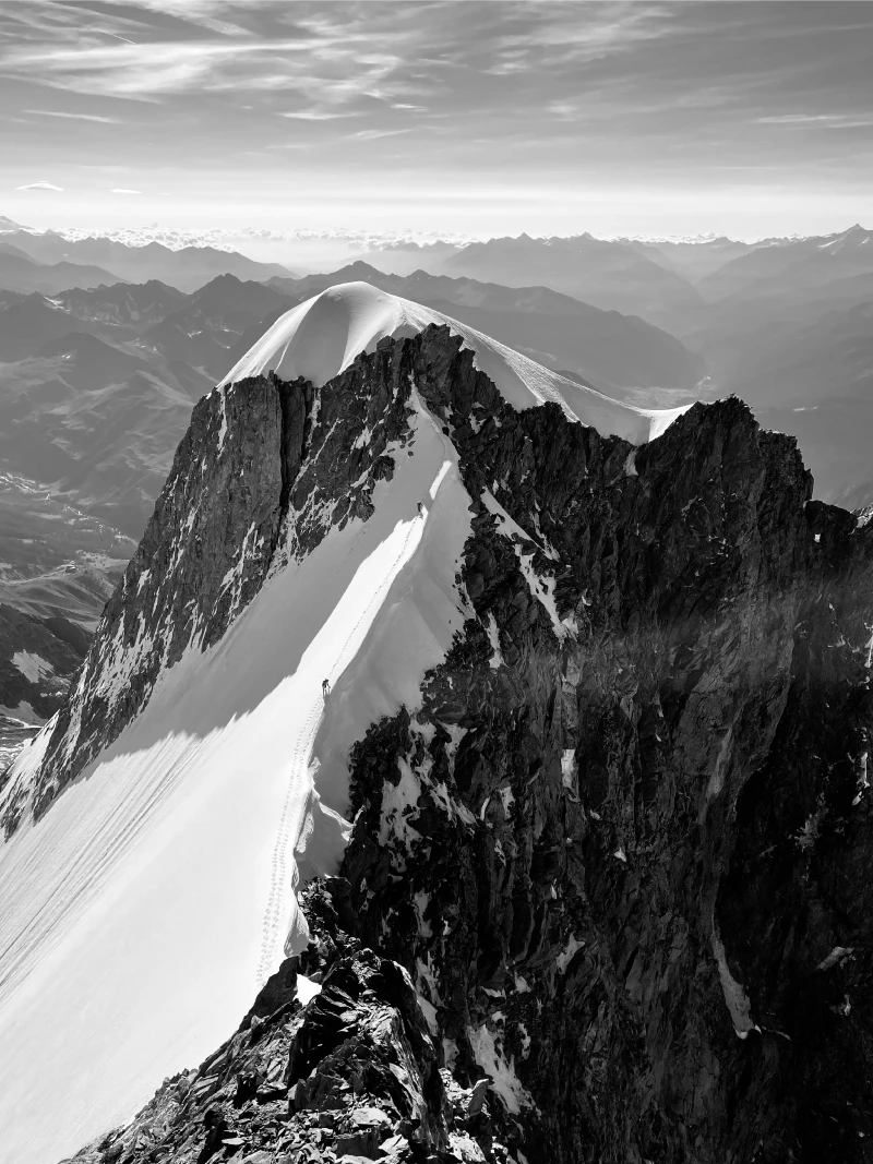 A black and white photograph of hikers walking along a snowy mountain ridge, taken from a distance.