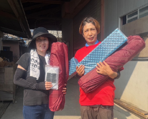 Two local women in a disaster-affected area holding relief supplies, including mats.