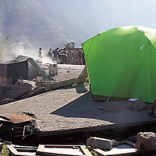 A scene of people living in evacuation, having set up Montbell tents. The surrounding area shows collapsed houses still remaining.