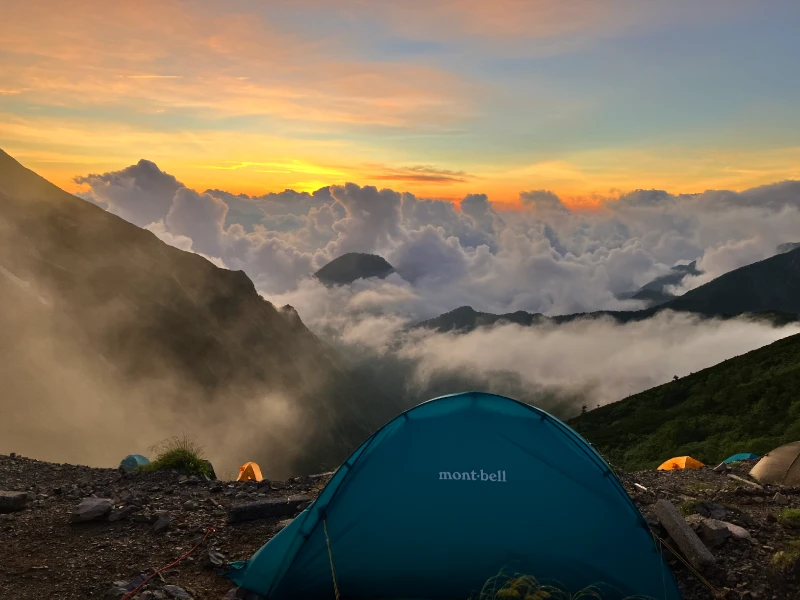 Multiple tents are set up at a camping site on a mountaintop, with a sea of clouds stretching behind them.