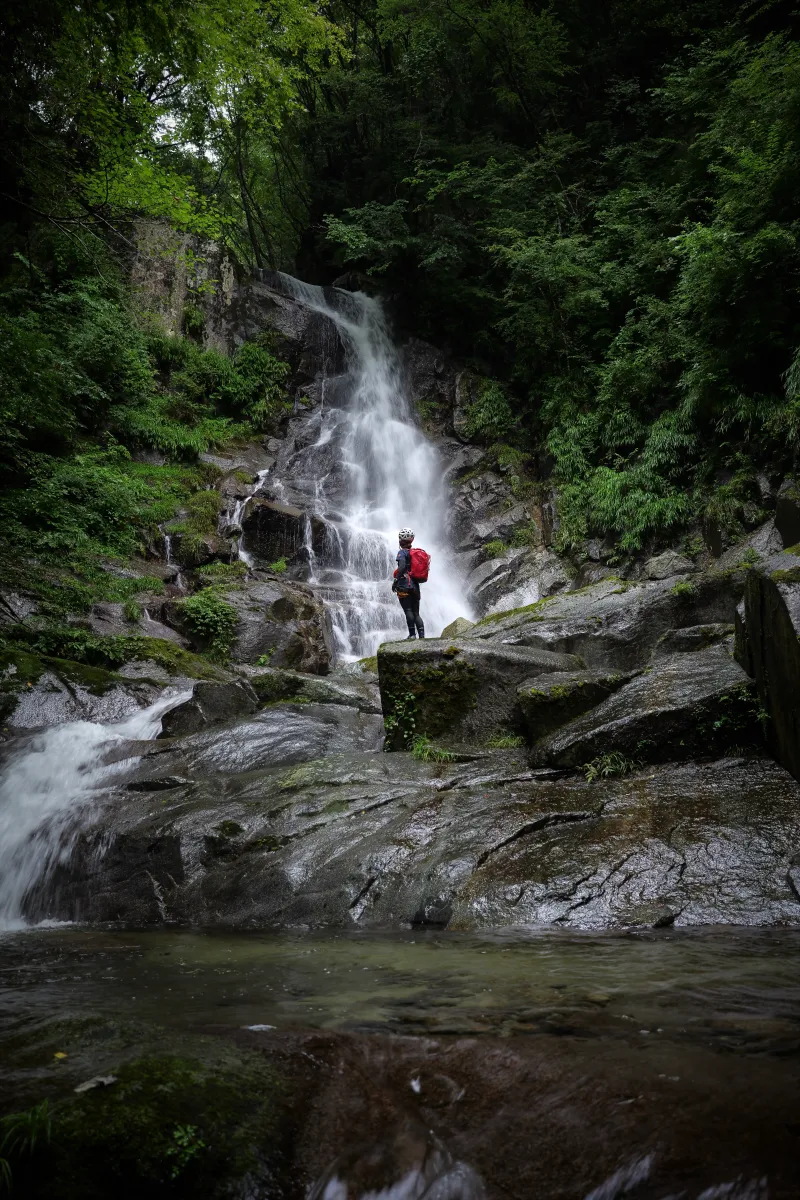 A person wearing a red backpack is seen from behind, looking up at an impressive 30-meter waterfall.