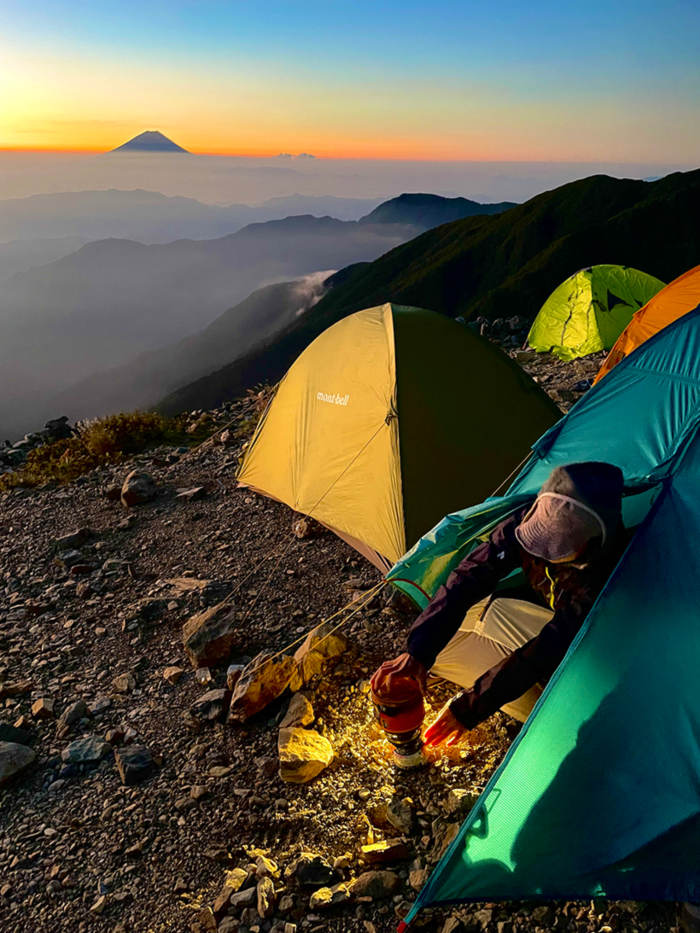 In the morning glow, a man emerges from a blue tent, his upper body visible as he boils water using a Jetboil. Beyond him stretches a sea of clouds, with the silhouette of Mount Fuji visible in the distance.