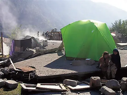 A scene of people living in evacuation, having set up Montbell tents. The surrounding area shows collapsed houses still remaining.